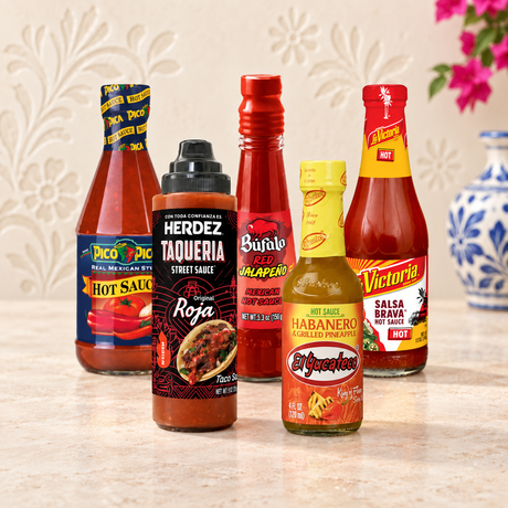 Five hot sauce bottles arranged together on a light stone surface against a beige patterned wall. The bottles include Ticonderica, Herdez Taqueria Street Sauce, Buffalo Red Pepper Sauce, El Vigor Habanero Hot Sauce, and Victoria Salsa Brava.